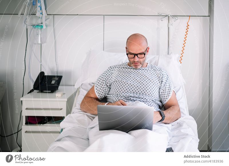 Serious male patient working on laptop on ward bed man serious focus workaholic hospital device clinic watch professional concentrate digital medicine sick ill