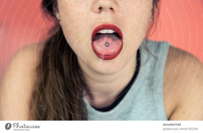 Close up portrait of young woman with freckles sticking out pierced tongue, showing her tongue piercing with pink background fashion person girl erotic face eye