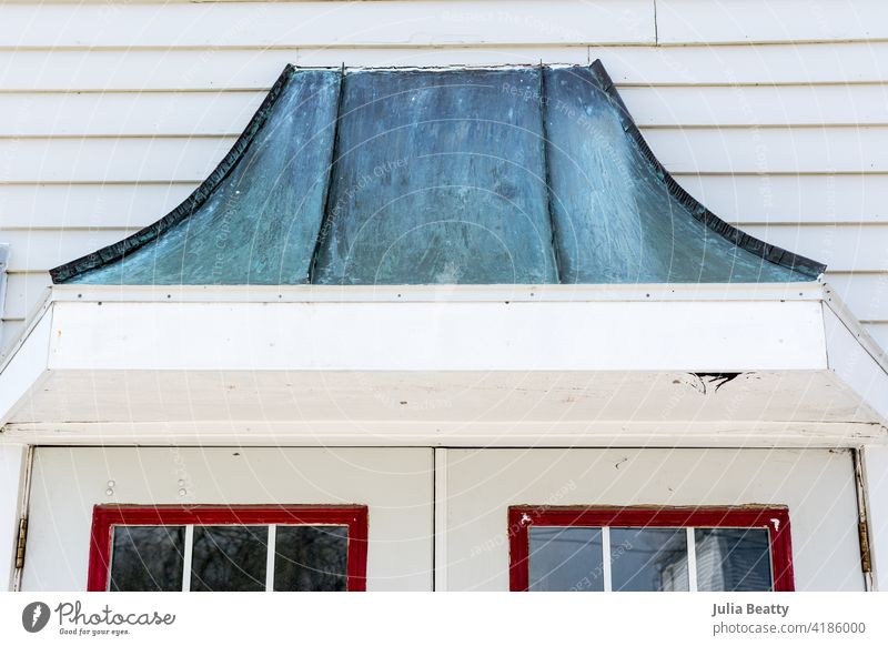 Metal gable with turquoise patina over a set of French doors with red trim; old facade of a small town building dormer disrepair fix damaged wood siding metal