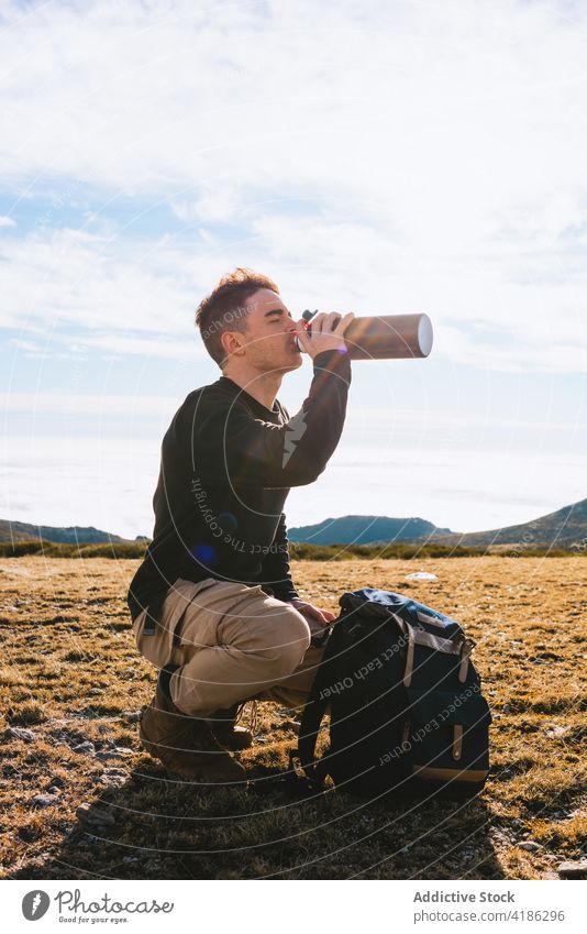 Young male hiker enjoying hot drink while sitting on haunches at hillside man beverage thermos traveler trip relax eyes closed mountain nature young casual