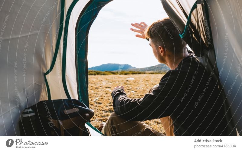 Young pensive camper relaxing in tent in sunny morning man rest dreamy traveler mountain admire trip journey recreation wanderlust male young casual style