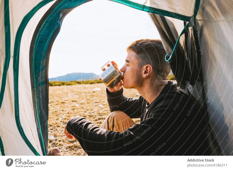 Stylish young guy enjoying hot drink during camping in mountainous countryside man beverage tent recreation hiker confident style nature grassland male trendy