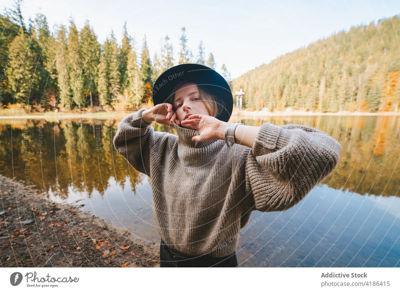 Stylish traveler in hat on shore against lake tourist cool style trip reflection tree woman sky stylish apparel garment confident trendy water mirror beautiful