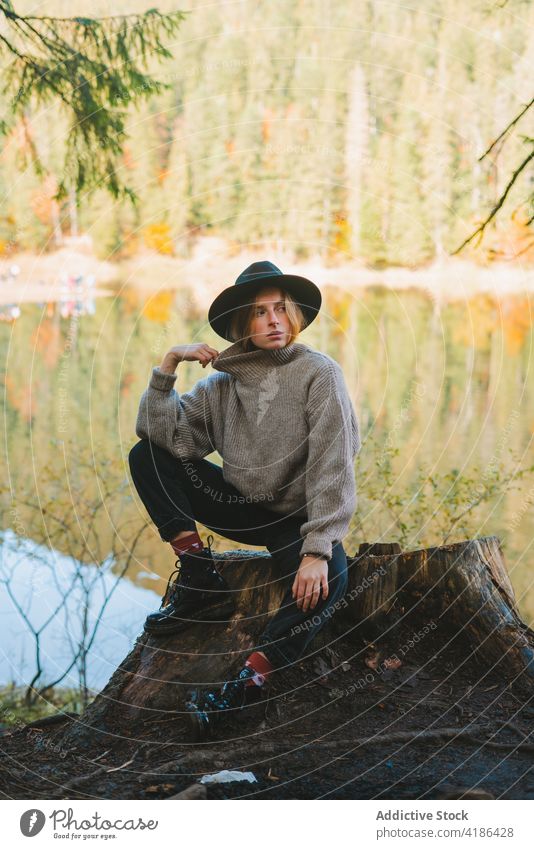 Stylish traveler on stump against lake in autumn tourist touch face cool style rest journey tree woman reflection nature wear stylish trip vacation casual sit