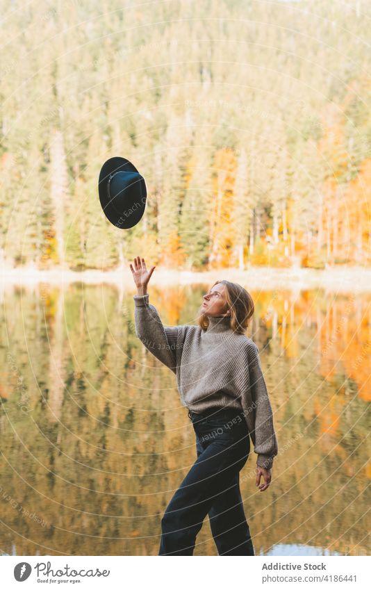 Woman throwing hat in air while walking against lake woman tree reflection nature attentive traveler casual style trip outfit garment knitted sweater tourist
