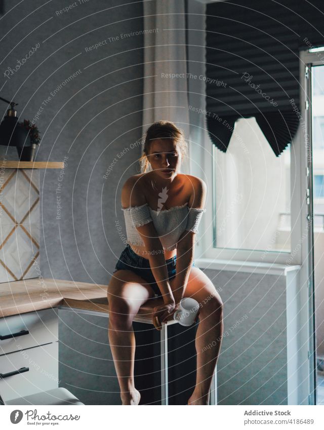 Pretty woman sitting on kitchen counter unemotional calm dreamy casual thoughtful feminine mug drink tranquil charming bare shoulders attractive beautiful