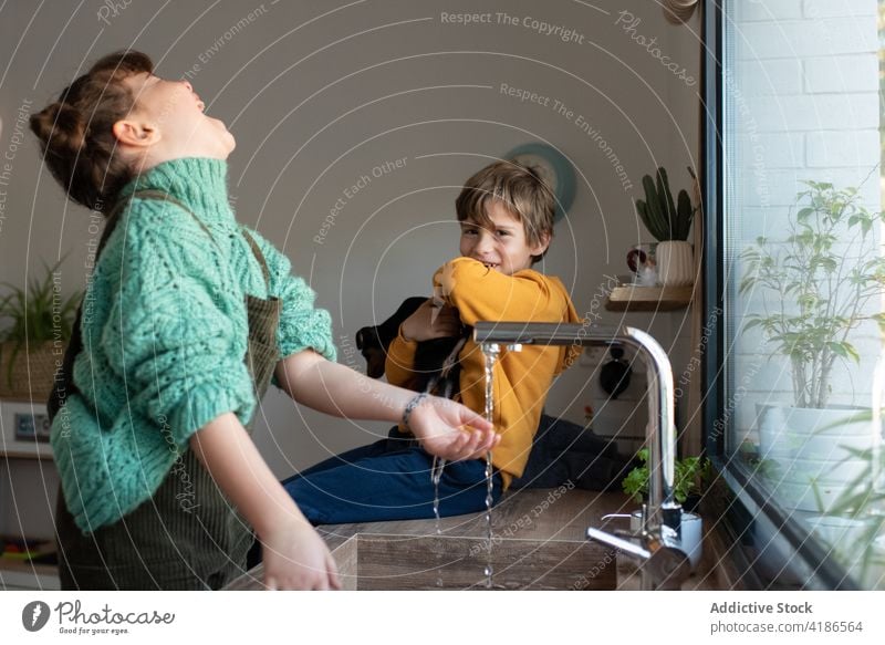 Delighted children playing in kitchen near sink sibling water together having fun delight carefree home weekend joy sister brother kid game adorable domestic