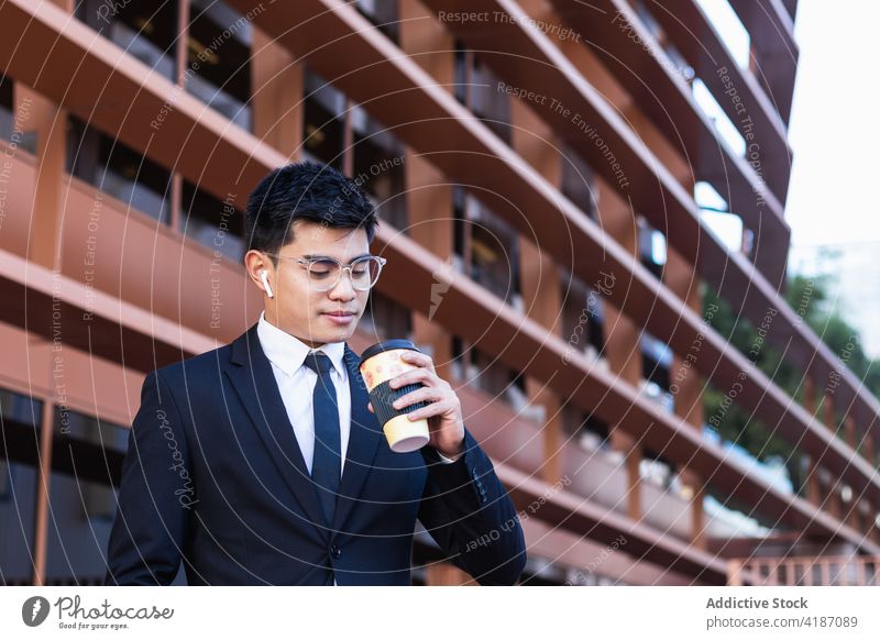 Handsome ethnic businessman with takeaway coffee in city handsome suit entrepreneur to go masculine street male asian wireless earbuds beverage drink paper cup