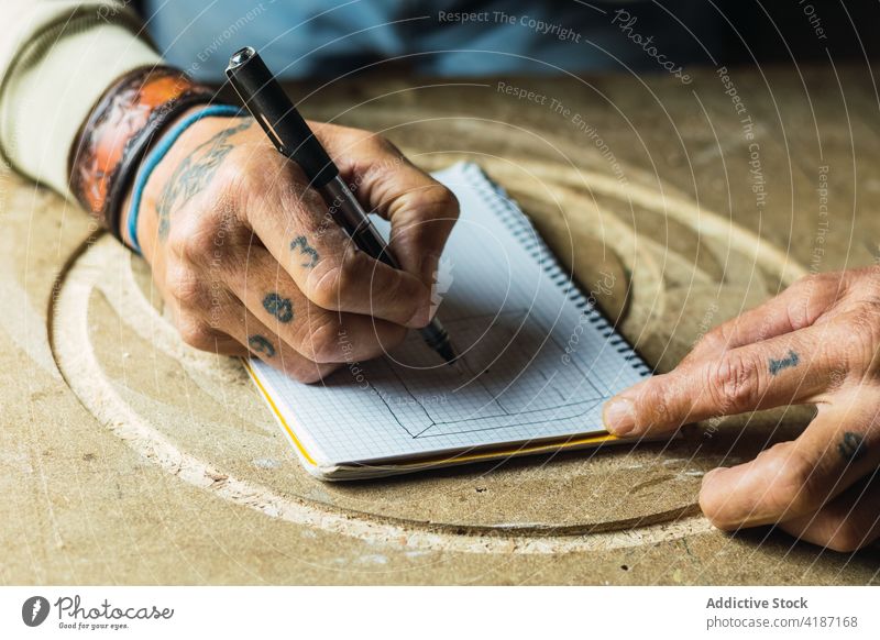 Crop woodworker drawing sketch in notebook carpentry draft carpenter mature man joinery male middle age tattoo job workshop occupation workbench hobby craft