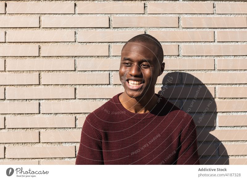 Happy young African American male standing near billet wall man casual happy style masculine ponder shadow brick wall town alone black knitted garment denim