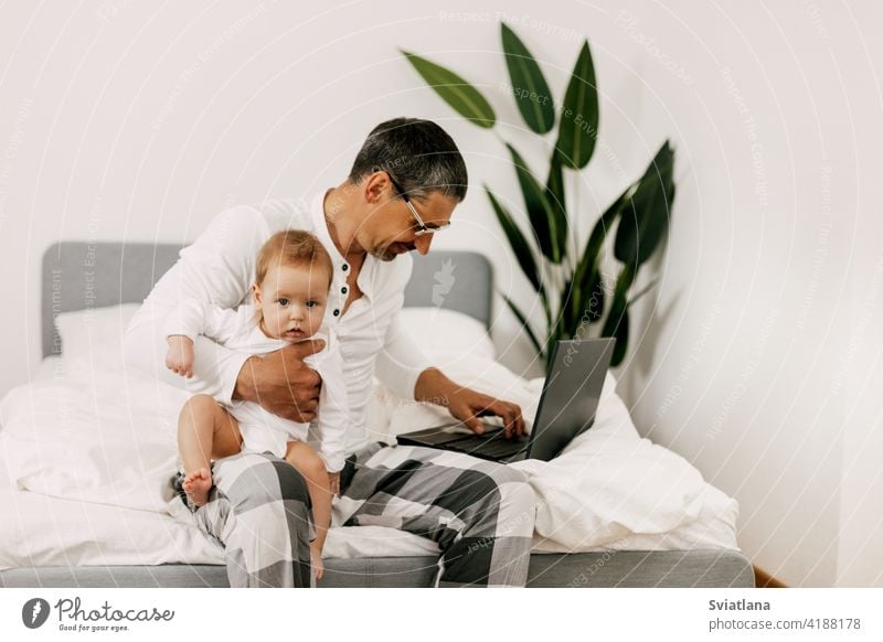 Dad sits on the bed, works on a laptop at home with a baby on his lap, smiles and looks at the screen together Home internet dad home office son remote work