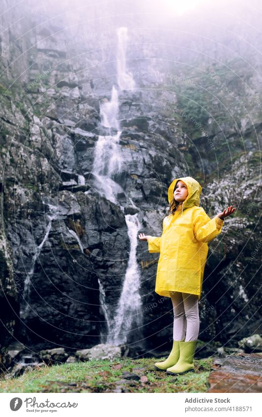 Girl in yellow raincoat against waterfall in mountains girl arms raised tourism highland nature wanderlust travel dreamy cascade fast flow bright energy dynamic
