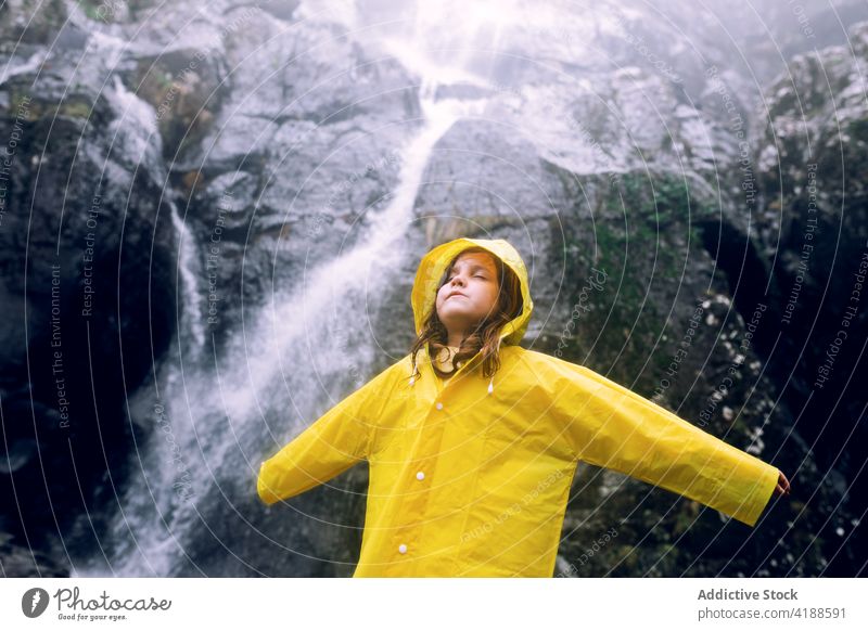 Girl in yellow raincoat against waterfall in mountains girl arms raised tourism highland nature wanderlust travel dreamy cascade fast eyes closed flow bright