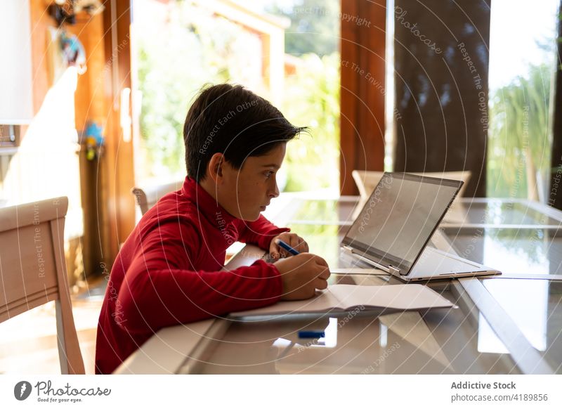 Boy doing homework at table with laptop child write notebook boy diligent study schoolboy pupil handwriting device smart gadget education task knowledge sit