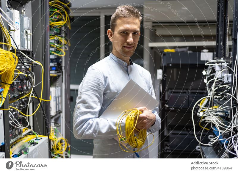 Smiling technician with laptop and coil of electrical wires man cable electronic engineering service facility male network server data positive center