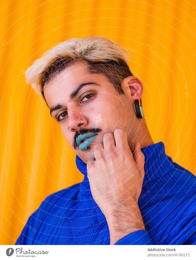 Stylish man with makeup looking at camera androgynous trendy model style fashion alternative apparel visage male blue lip lipstick emotionless outfit yellow