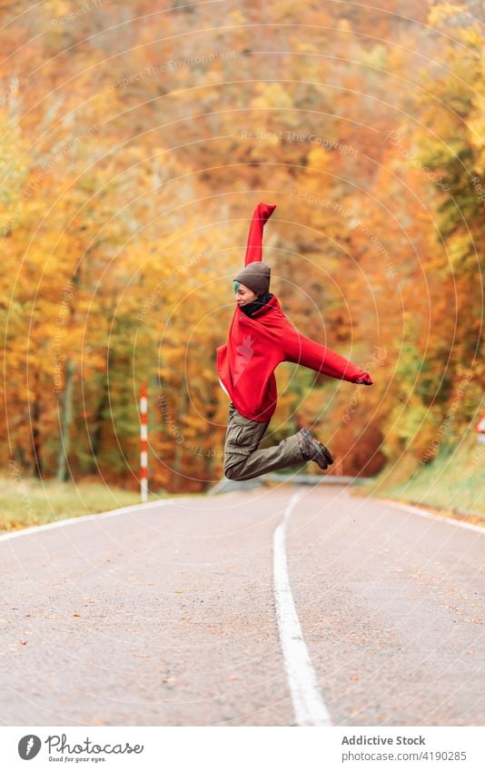 Happy woman in outerwear jumping on rural road in autumn excited arms raised forest mid air freedom joy adventure wanderlust above ground energy season traveler