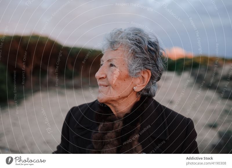 Positive senior woman smiling while resting on sandy seashore in evening smile relax beach tourist happy portrait holiday glad coast cloudy sky female elderly