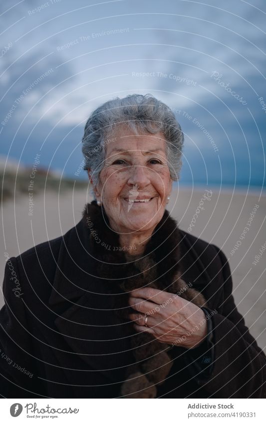 Positive senior woman smiling while resting on sandy seashore in evening smile relax beach tourist happy portrait holiday glad coast cloudy sky female elderly