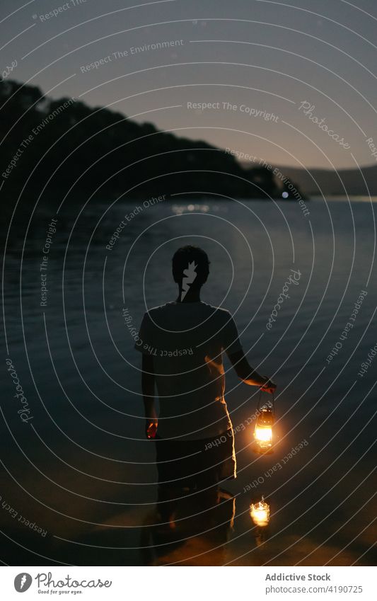 Anonymous traveler with lantern admiring lake against mountain at night admire nature landscape sky man shiny ripple water silhouette tourist contemplate
