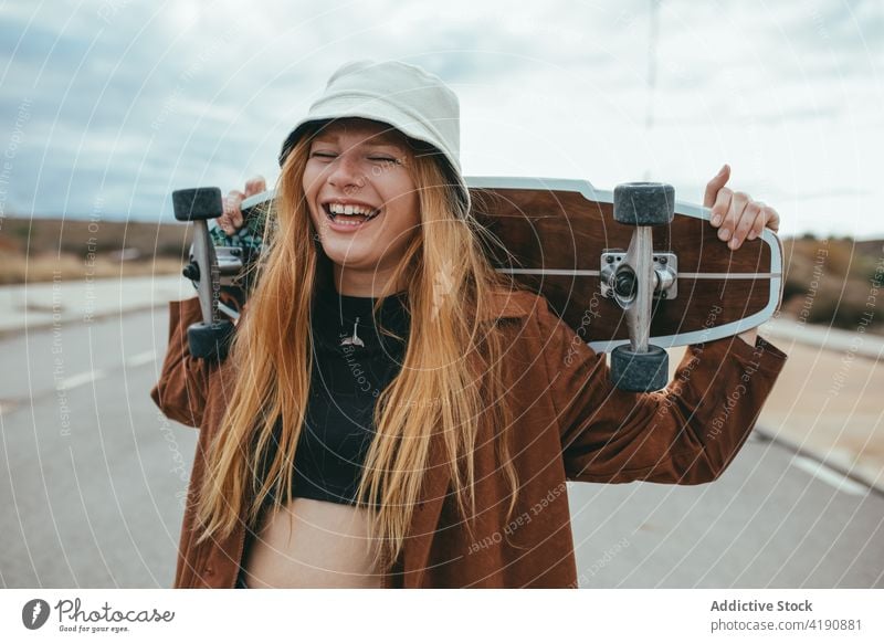 Joyful young woman laughing while standing on road with skateboard in hands eyes closed longboard activity self assured cheerful cool delight positive happy