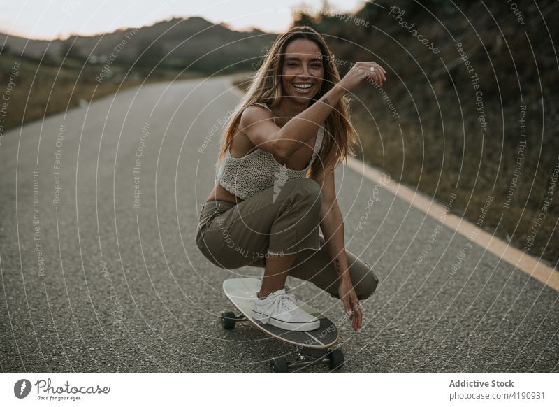 Cheerful female skater hunkering down on cruiser board on rural road woman carefree cheerful activity ride haunch toothy smile countryside twilight evening