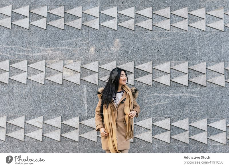 Smiling Asian woman in coat and scarf standing near building positive individuality ornament wall appearance geometry smile carefree female cheerful optimist