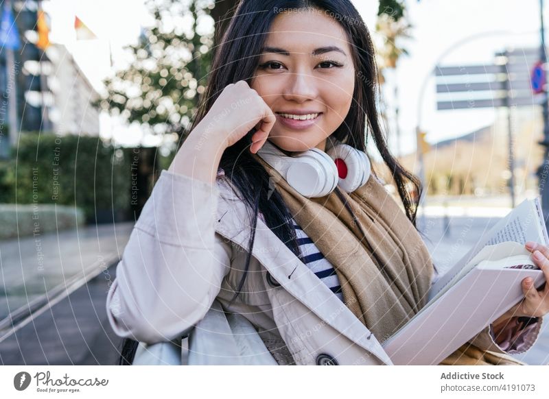 Smiling Asian woman reading book sitting on bench in park optimist story literature hobby headphones wireless smile female fiction knowledge toothy smile casual