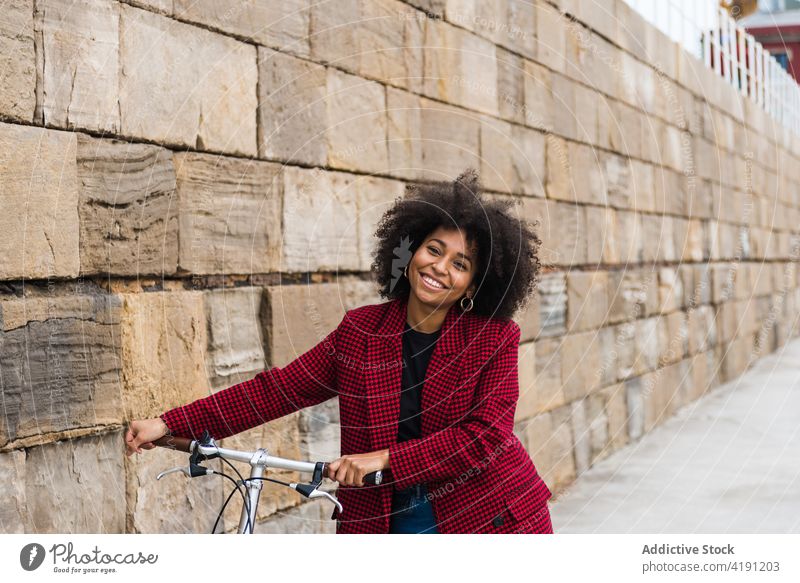 Delighted ethnic woman walking with bicycle in city street bike cheerful smile urban vehicle female black african american old stone wall building stroll enjoy