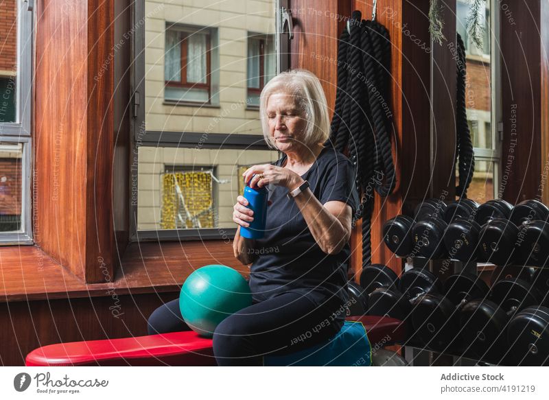 Elderly sportswoman opening bottle of water during training in gym athlete break workout equipment gymnasium exercise ball small bench rest sportswear pilates