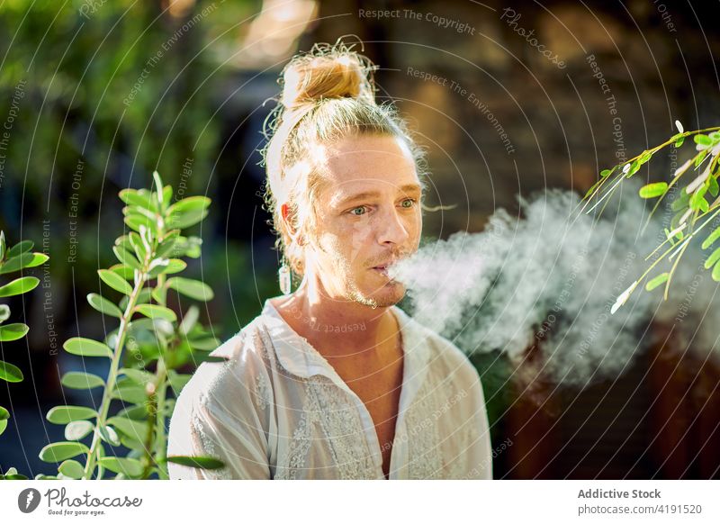 Hipster blond man smoking cigarette in garden male hipster handsome style self assured outdoors summer harmony idyllic calm tranquil hippie serene peaceful