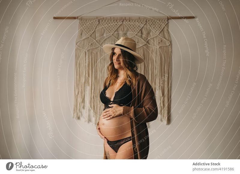 Pregnant woman touching belly and looking away pregnant smiling happy pensive style touch belly motherhood prenatal expect apartment lingerie female hat