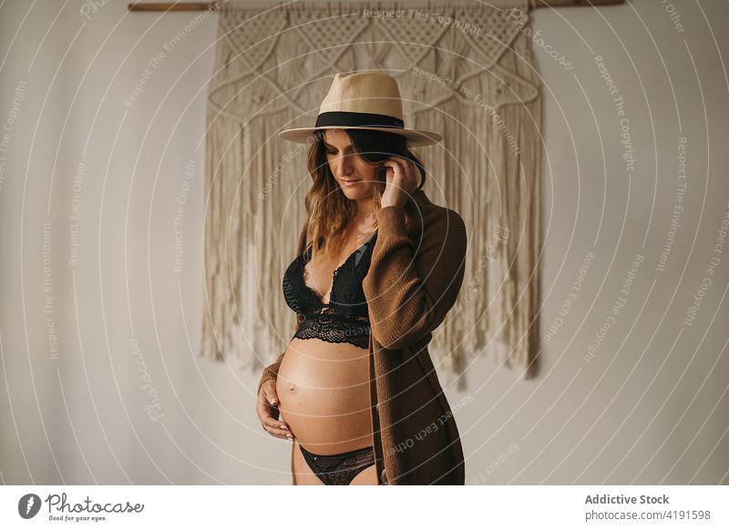 Pregnant woman touching belly and looking down pregnant pensive style touch belly motherhood prenatal expect apartment lingerie female hat parenthood dreamy