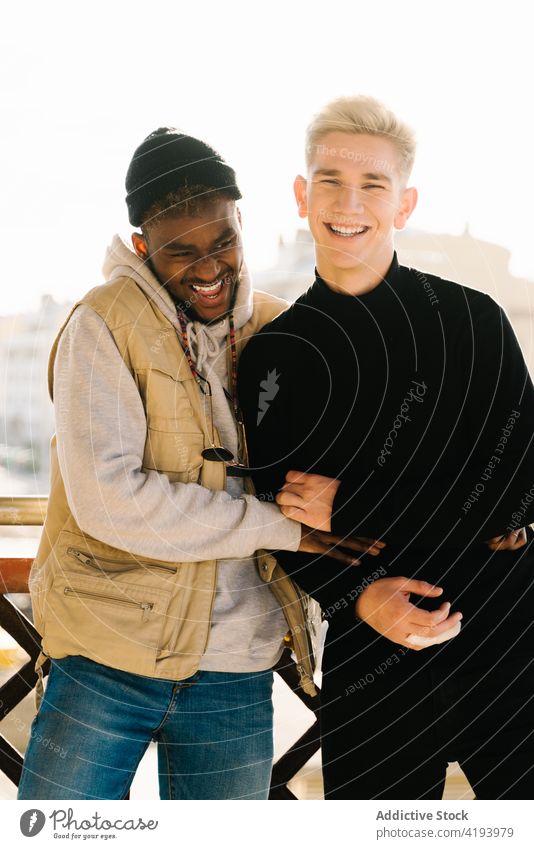 Cheerful young diverse boyfriends laughing and cuddling on balcony couple gay smile hug homosexual lgbt terrace together love spend time cheerful trendy style