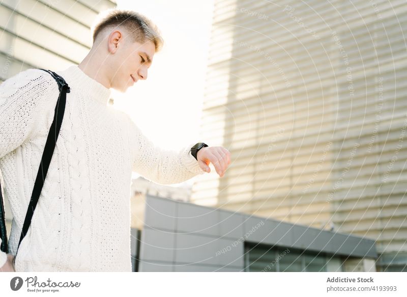 Trendy young man checking time on wristwatch while standing near modern buildings street city wait appointment punctual architecture downtown urban male trendy