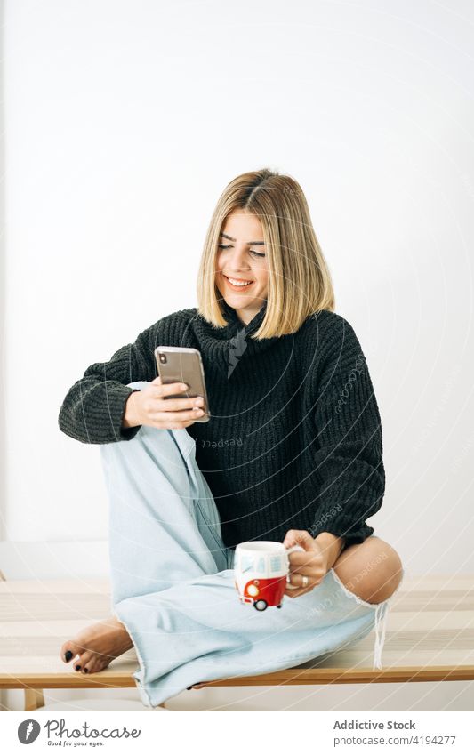 Smiling woman with hot drink chatting on smartphone at home internet online smile cozy using gadget cellphone device cup coffee beverage tea comfort rest relax