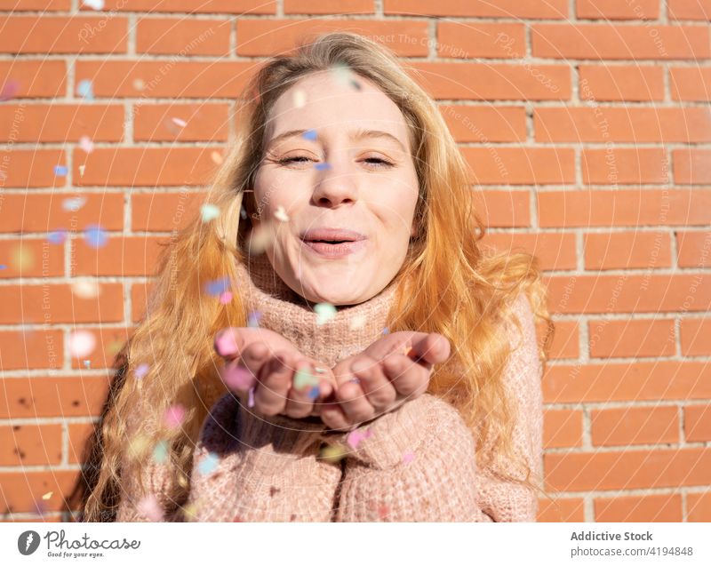 Delighted young lady blowing confetti on street woman smile happy delight brick wall having fun glad holiday optimist cheerful joy female long hair wavy hair