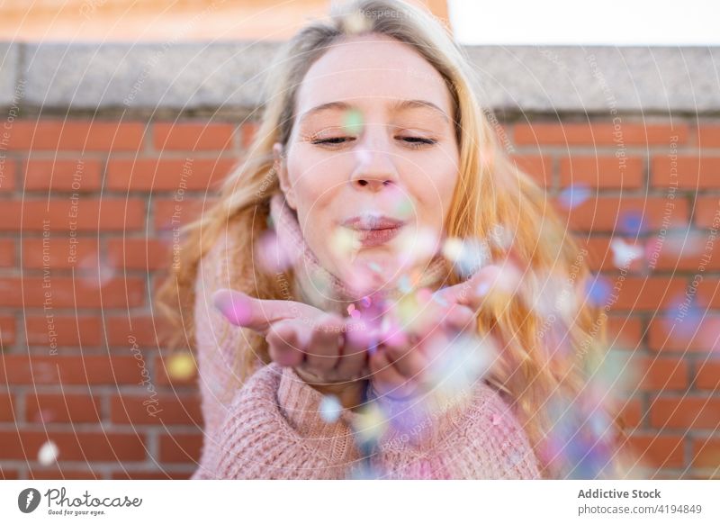 Delighted young lady blowing confetti on street woman smile happy delight brick wall having fun glad holiday optimist cheerful joy female long hair wavy hair