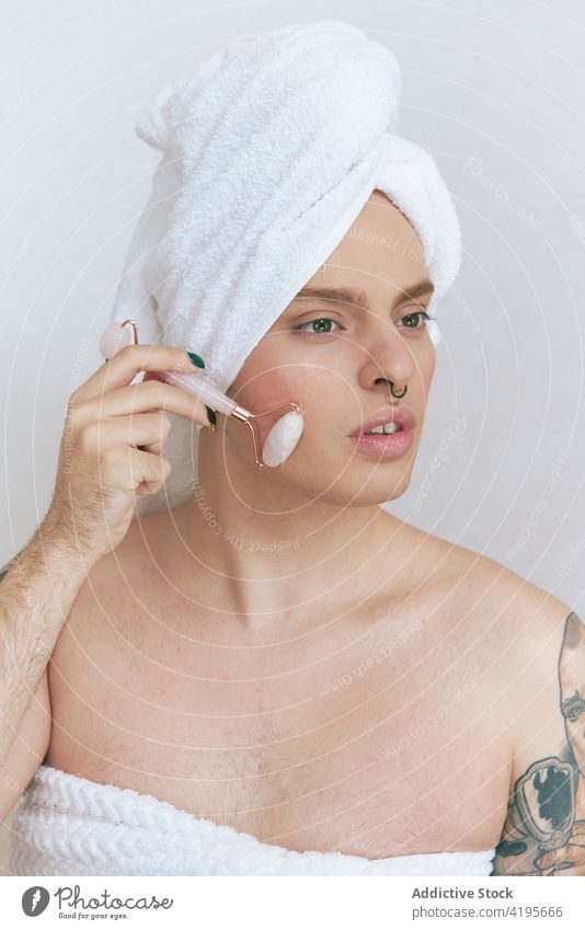 Queer massaging face with roller on white background queer massage facial beauty natural tattoo tender contemplative portrait man androgynous identity dreamy