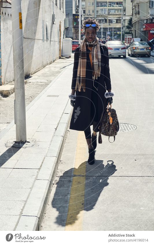 Stylish black woman walking on city road with shadow stylish fashion confident feminine accessory street style friendly sincere town building fur