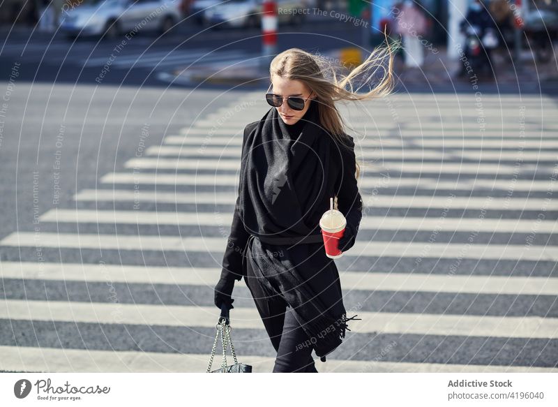 Stylish woman with refreshing drink to go crossing urban road stylish flying hair stroll fashion town lifestyle cup disposable plastic straw sunglasses