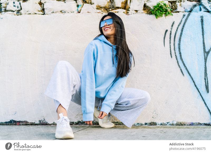 Stylish smiling woman in sunglasses on urban pavement fashion style individuality cool contemporary smile city squat hoodie trousers sneakers modern cheerful