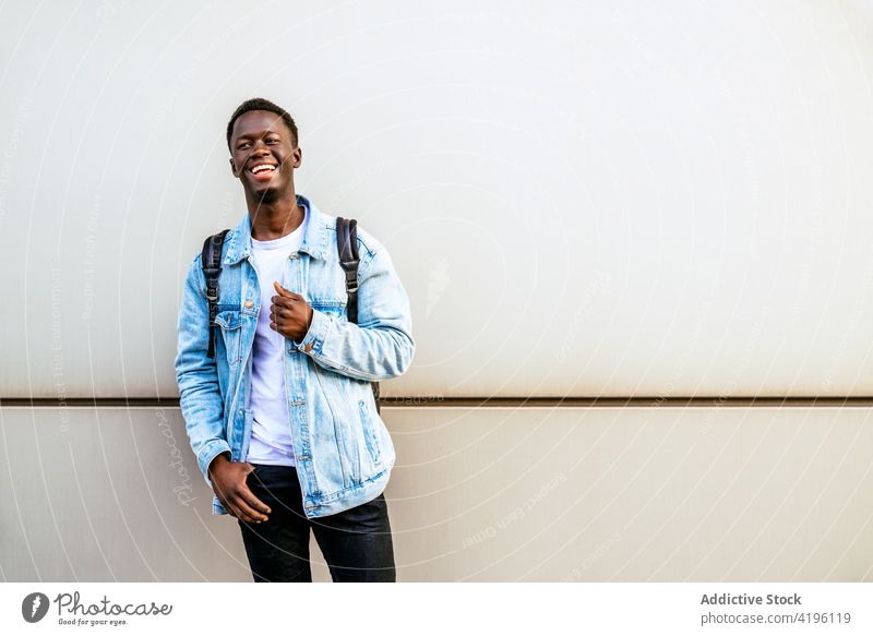 Smiling African American man with hand on hip near wall cheerful sincere casual style friendly happy enjoy denim jacket masculine bright modern cloth smile