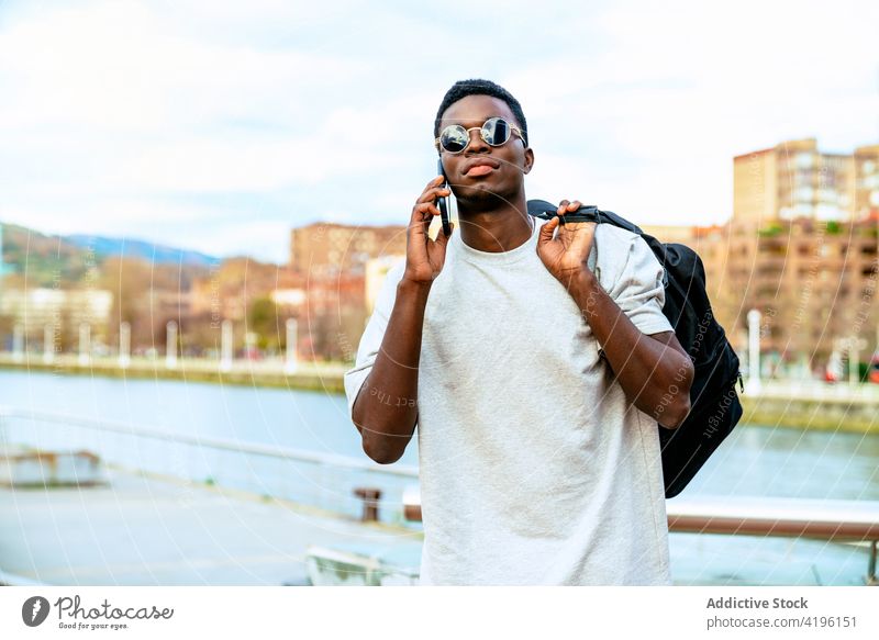 Black man in stylish sunglasses speaking on smartphone on embankment talk masculine macho trendy city using gadget phone call rucksack communicate serious cool