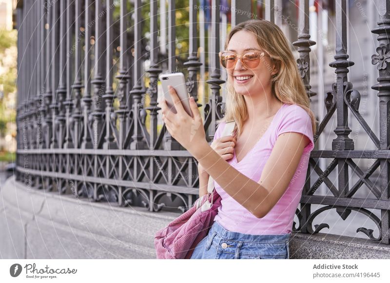 Smiling blonde teen woman surfing smartphone leaning on fence using teenage positive carefree browsing digital leisure internet female device mobile cellphone