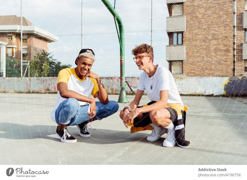 Two teenage boys squatting down and laughing on the urban basket court autumn black caucasian cheerful city colorful companionship daylight ethnic fellowship