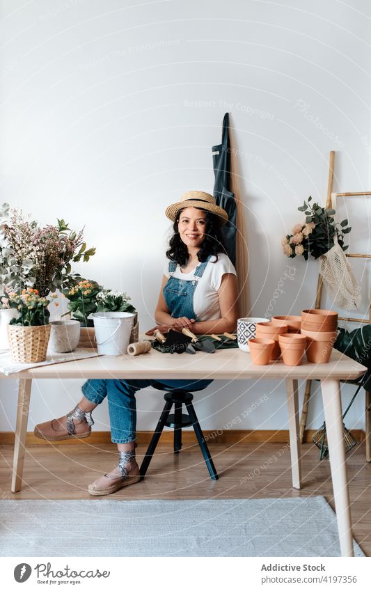 Gardener sitting at table in house near plant and flowers gardener cut foliage botany vegetate natural woman assorted tool trowel pot straw hat feminine