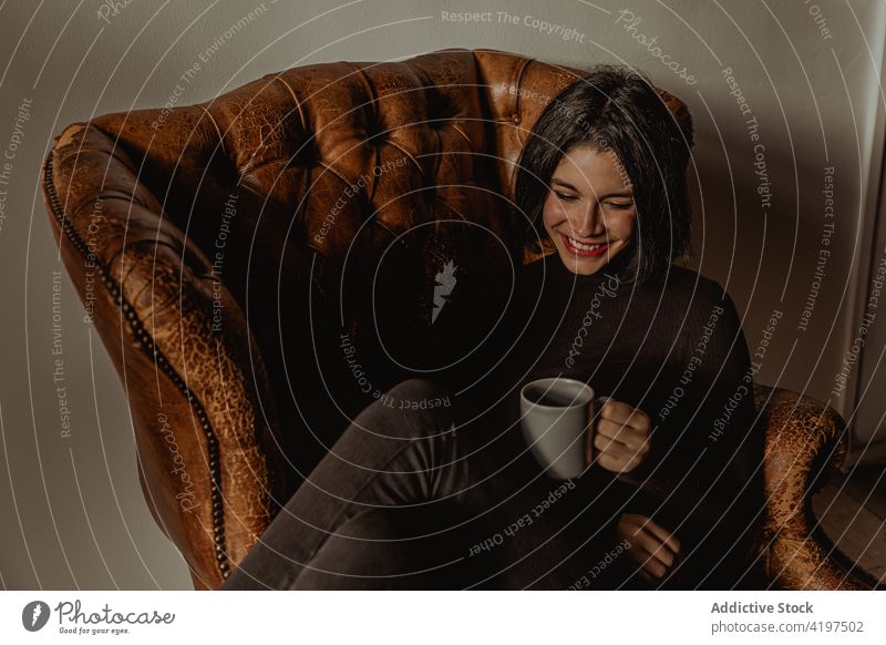 Cheerful woman with cup of drink in armchair cozy smile home enjoy comfort beverage delight female hot sit glad weekend young glee coffee tea lady shabby