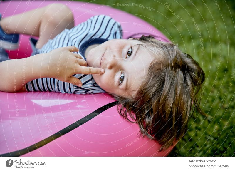 Playful kid lying on inflatable pink flamingo on lawn child park having fun playful joy vacation holiday childhood cheerful boy casual grassy nature glad