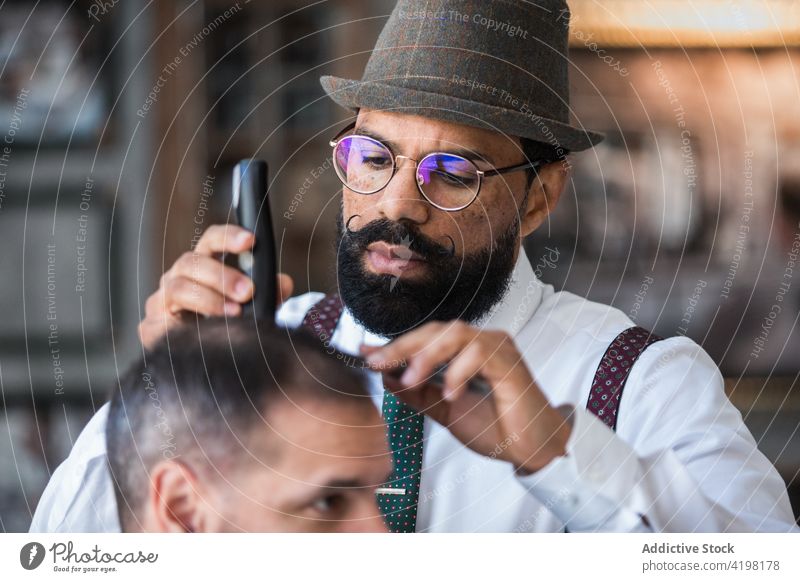 Ethnic hairdresser cutting hair of client with trimmer in barbershop clipper procedure beauty macho hipster men device dandy electric machine serious attentive